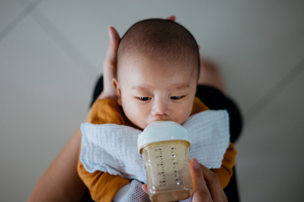 Parent cradling infant being fed formula with one hand behind head and other hand holding bottle